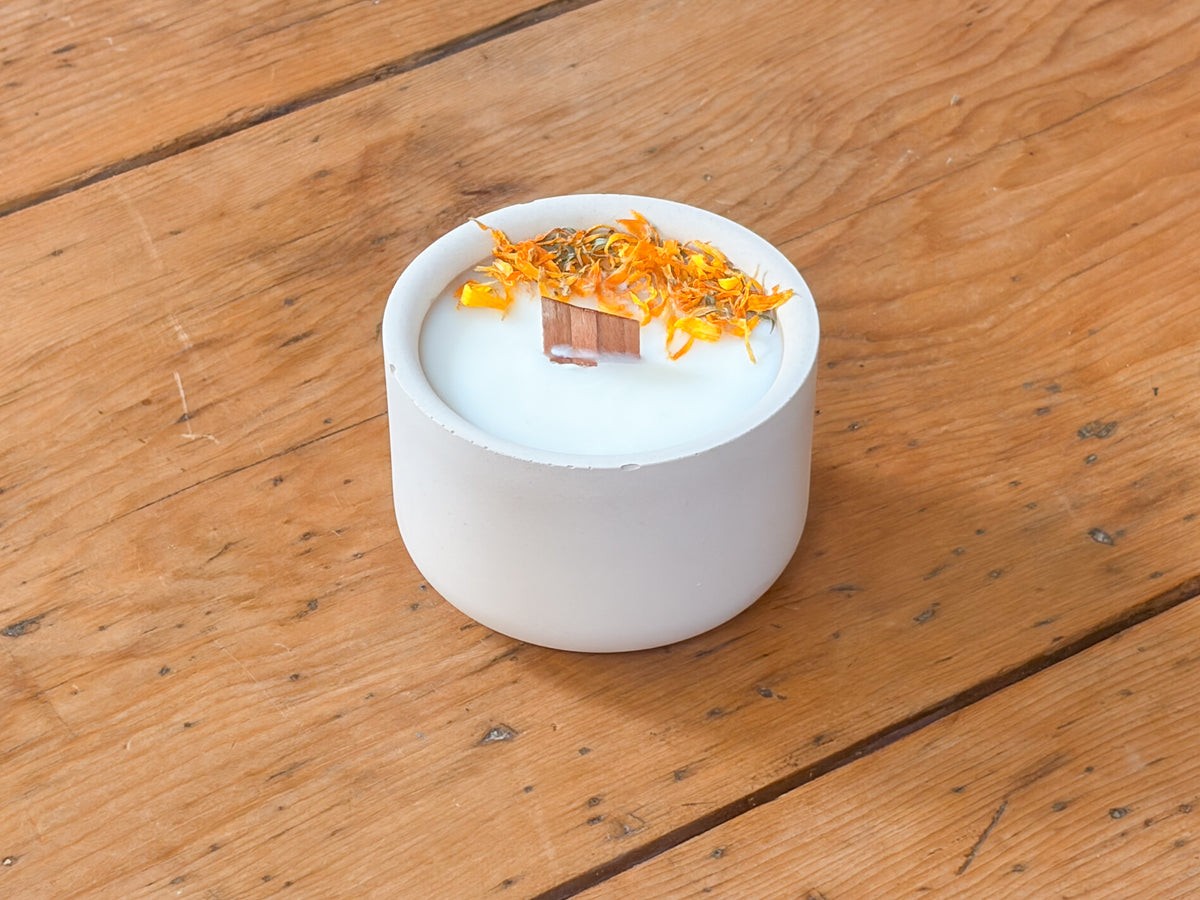A white ceramic candle jar filled with white wax, topped with dried orange flower petals and a small copper ingot, set on a rustic wooden plank surface.
