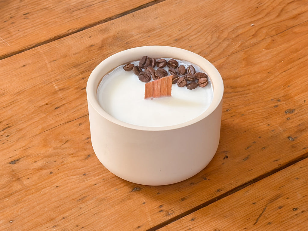 A ceramic soy candle in a round off-white pot, filled with white wax and topped with roasted coffee beans and a small wooden wick, set on a rustic wooden table.