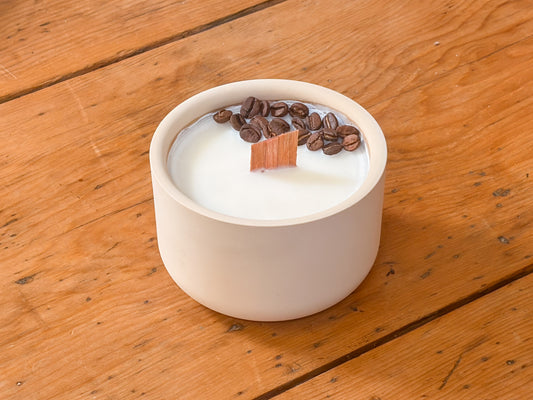 A ceramic soy candle in a round off-white pot, filled with white wax and topped with roasted coffee beans and a small wooden wick, set on a rustic wooden table.
