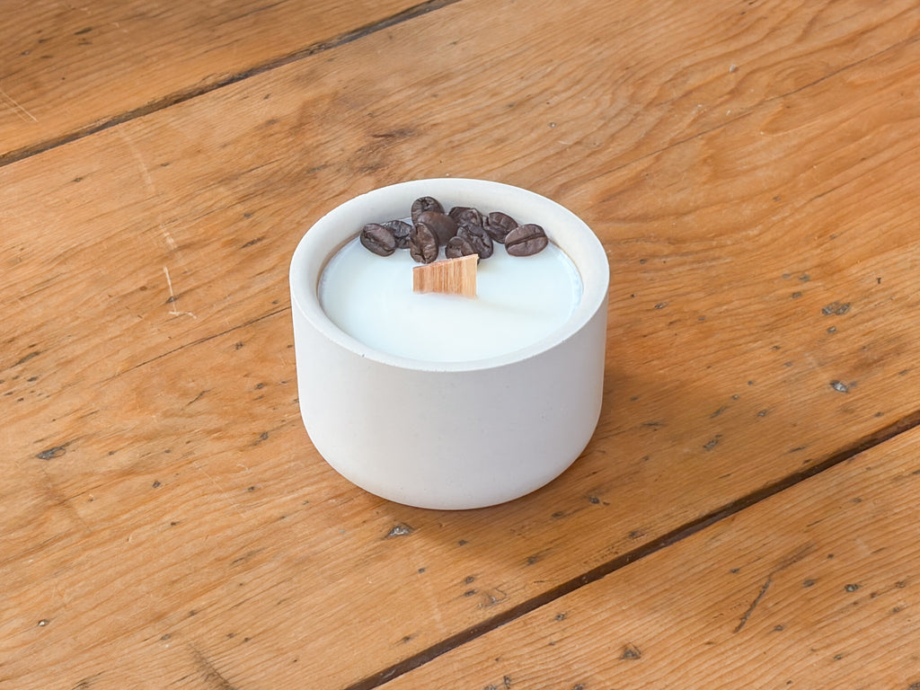 A white ceramic candle holder filled with white wax, topped with dark brown coffee beans and a small wooden wick, set on a rustic wooden table.