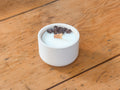 A white ceramic candle holder filled with white wax, topped with dark brown coffee beans and a small wooden wick, set on a rustic wooden table.