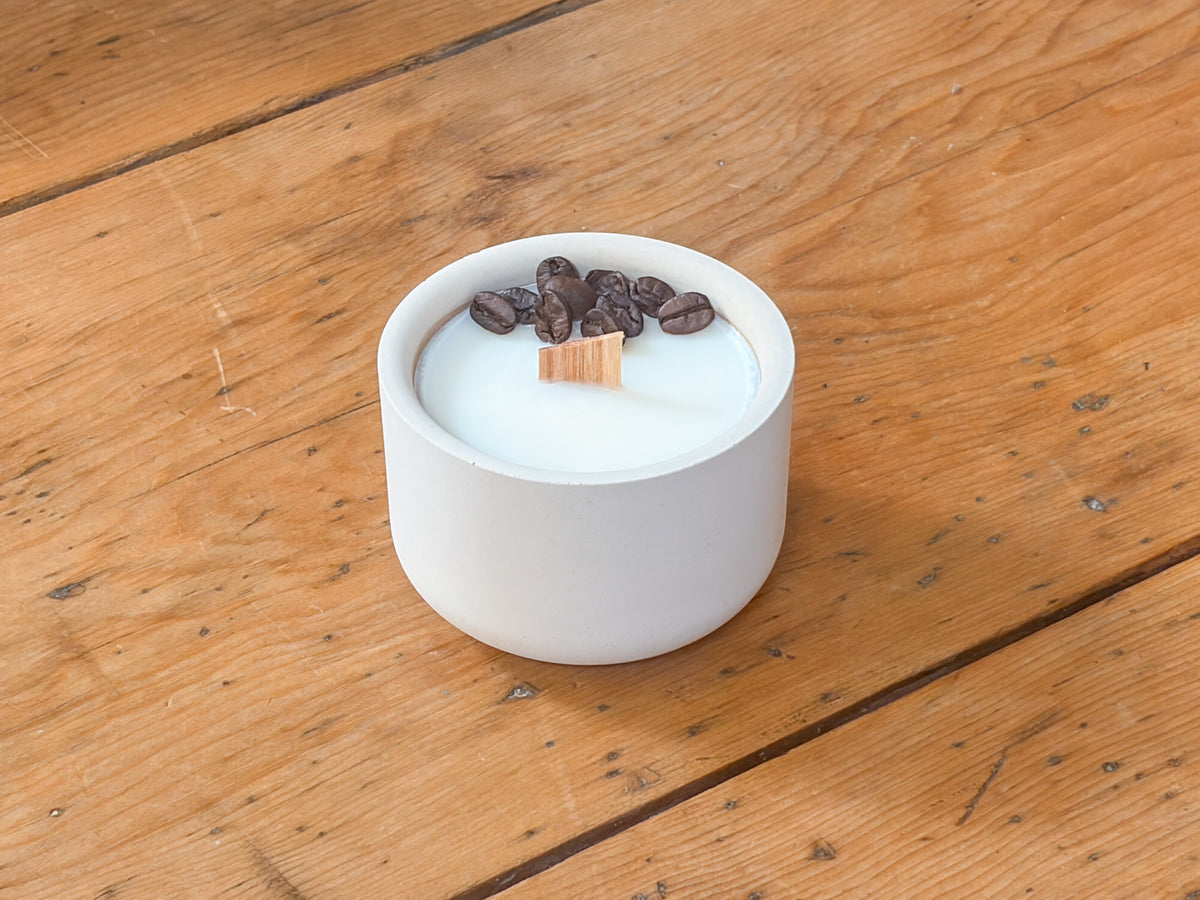 A white ceramic candle holder filled with white wax, topped with dark brown coffee beans and a small wooden wick, set on a rustic wooden table.