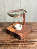A rustic wooden candle holder and tea light stand with a copper-colored metal ring and a clear glass top, set on a dark wood table against a neutral fabric backdrop.