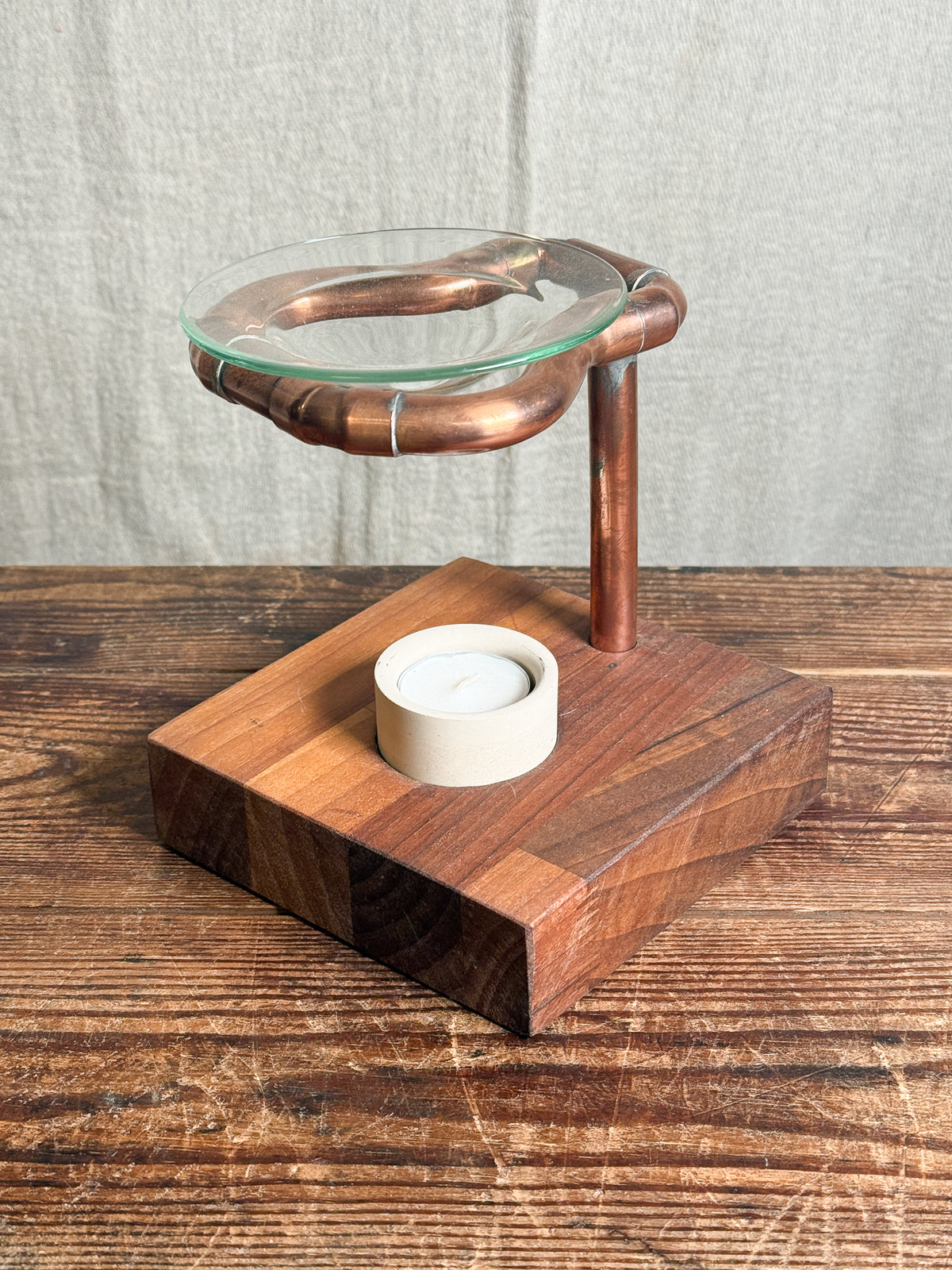A rustic wooden candle holder and tea light stand with a copper-colored metal ring and a clear glass top, set on a dark wood table against a neutral fabric backdrop.