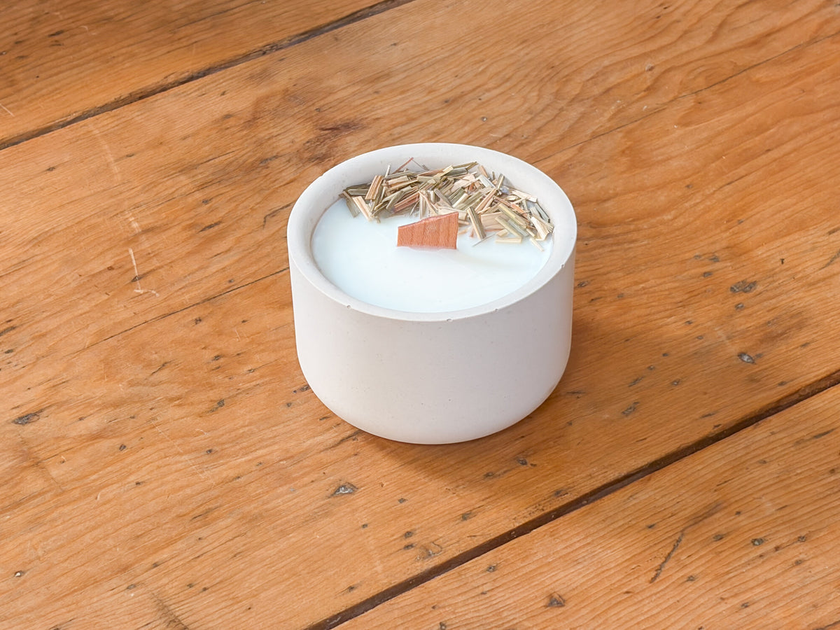 A small white ceramic candle holder filled with white wax and topped with dried botanicals, set on a rustic wooden surface.