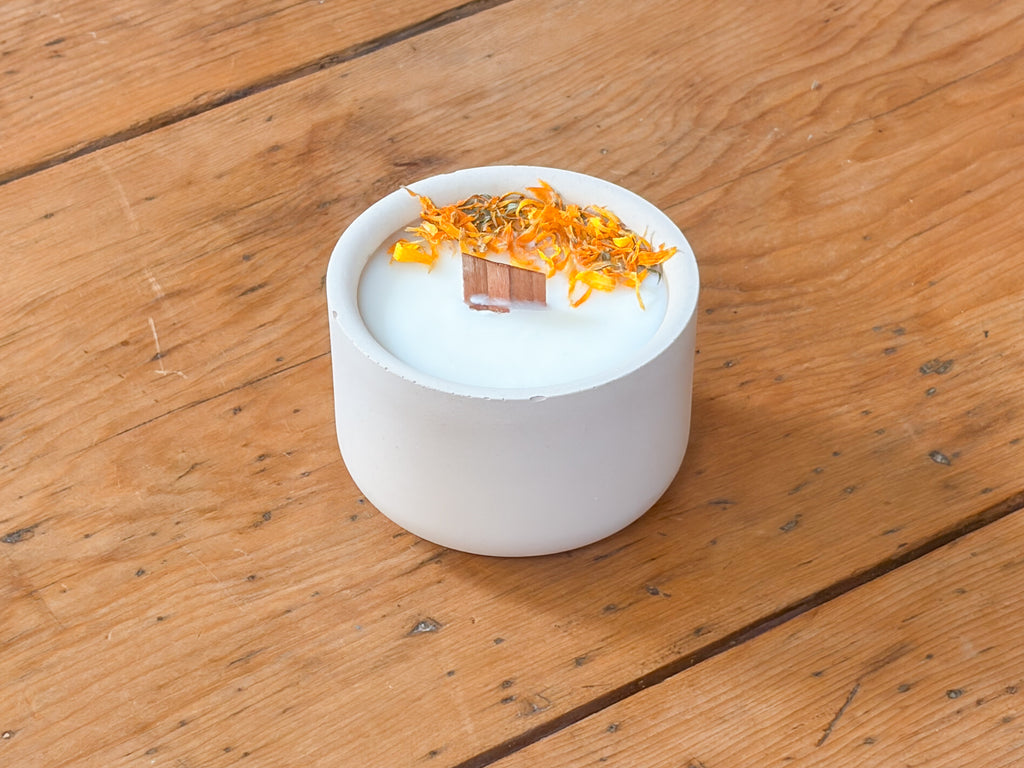 A white ceramic candle jar filled with white wax, topped with dried orange flower petals and a small copper ingot, set on a rustic wooden plank surface.