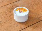 A white ceramic candle jar filled with white wax, topped with dried orange flower petals and a small copper ingot, set on a rustic wooden plank surface.