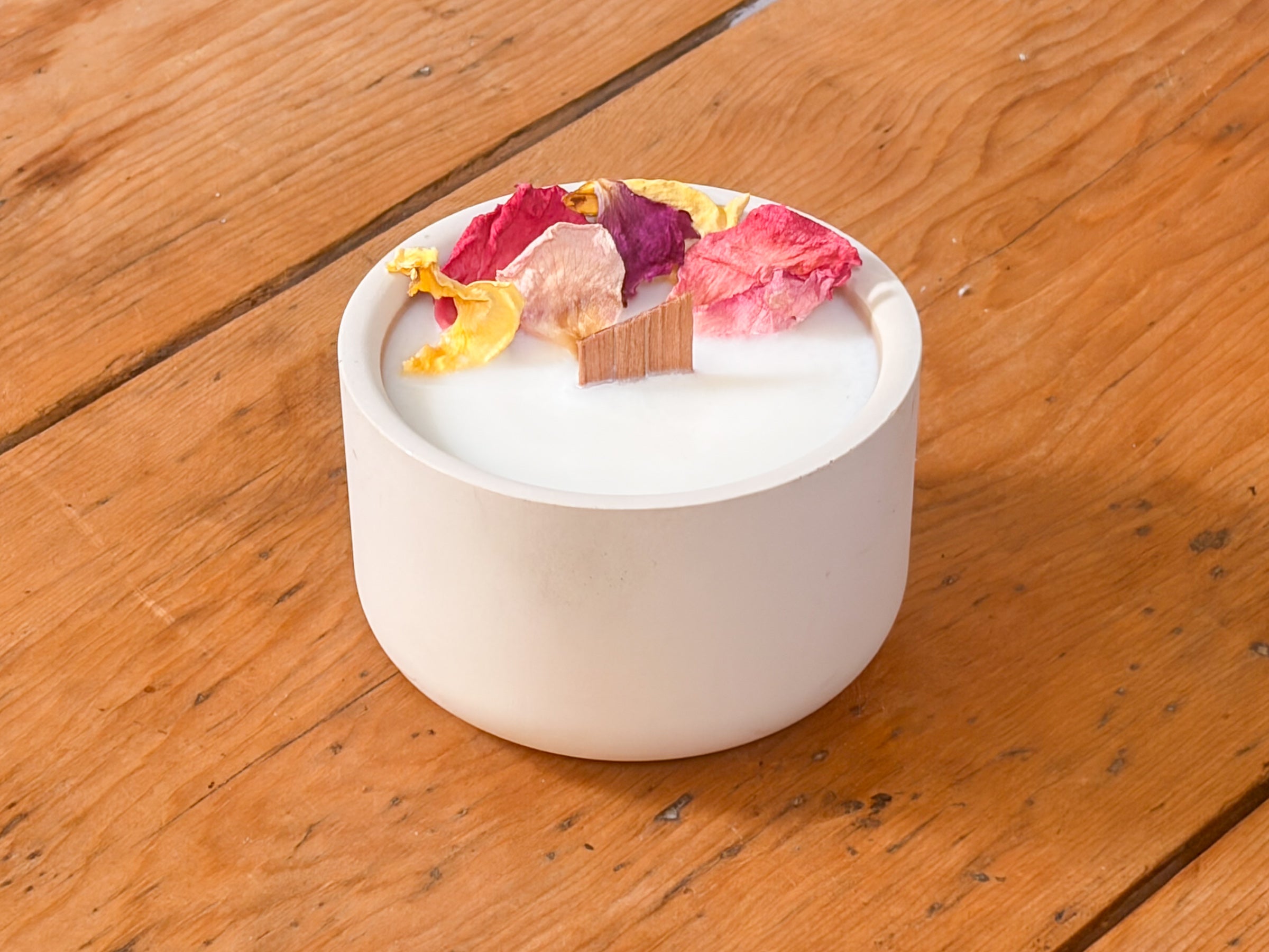 A white ceramic candle jar with dried flower petals on top, set on a rustic wooden table.