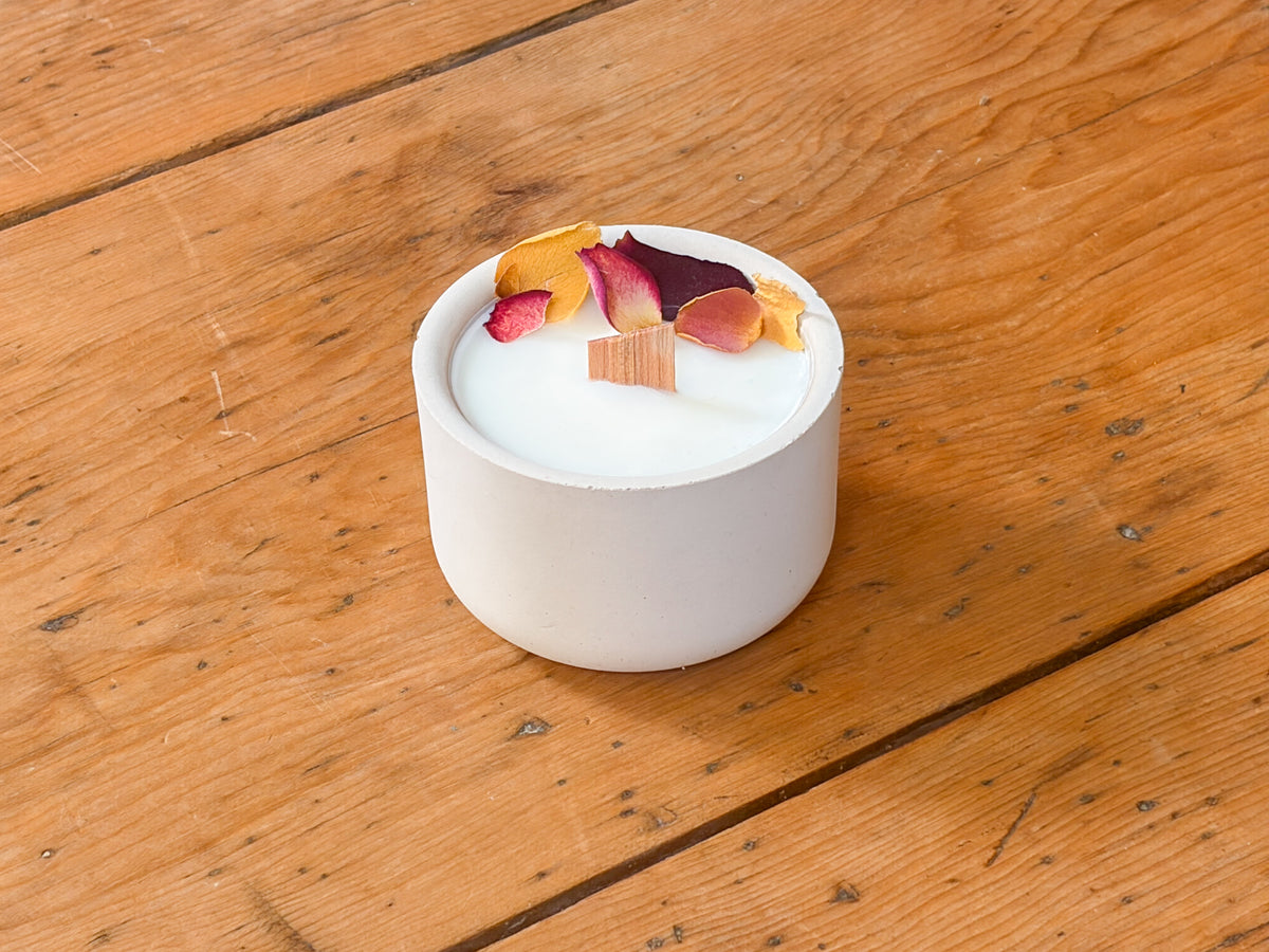 A white ceramic candle holder with white wax and dried flower petals on top, set on a rustic wooden table.