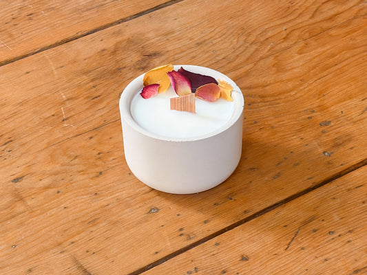 A white ceramic candle holder with white wax and dried flower petals on top, set on a rustic wooden table.