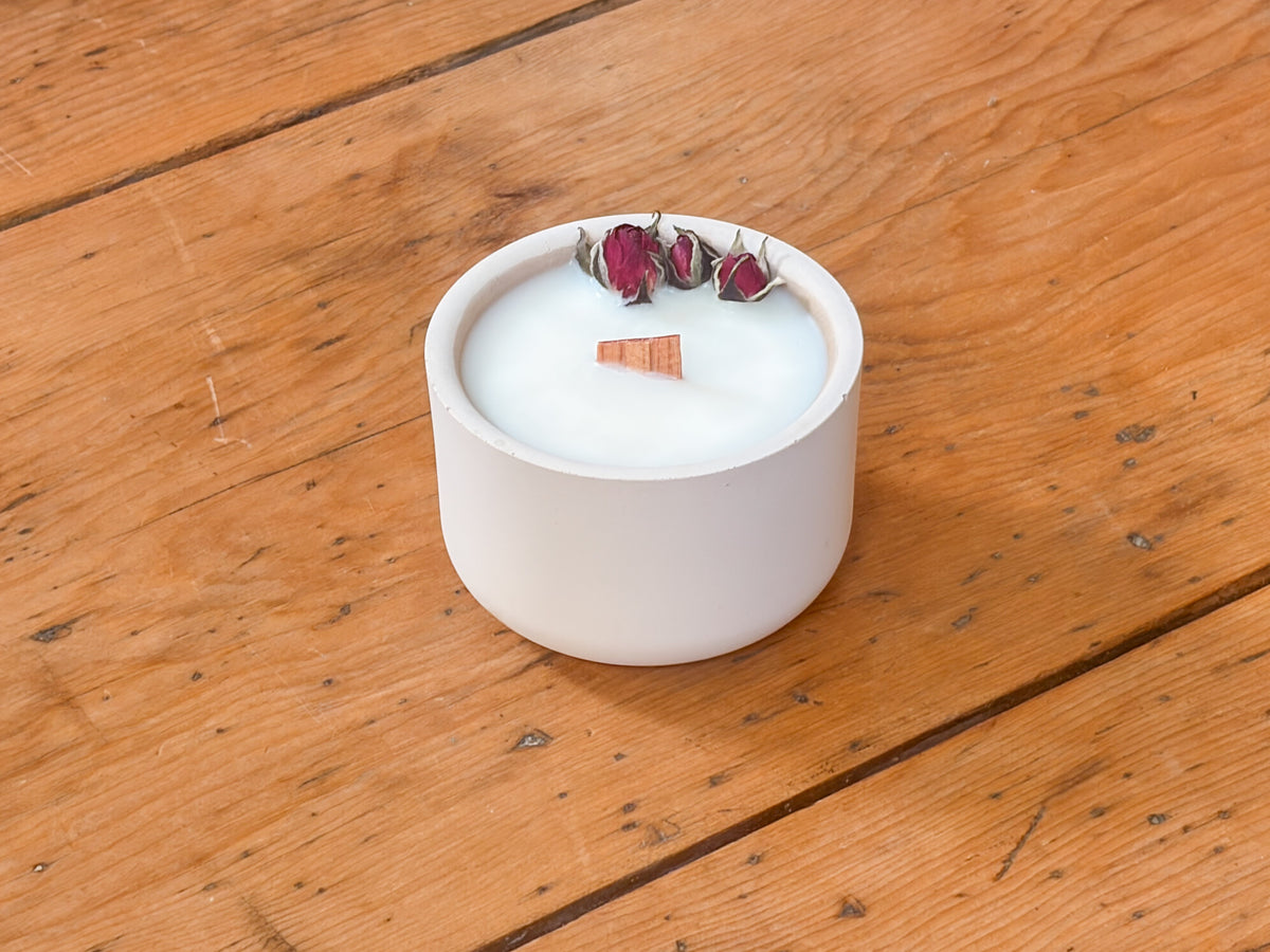 A white ceramic candle holder filled with white wax, decorated with three dried rose buds and a small cinnamon stick, set on a rustic wooden table.