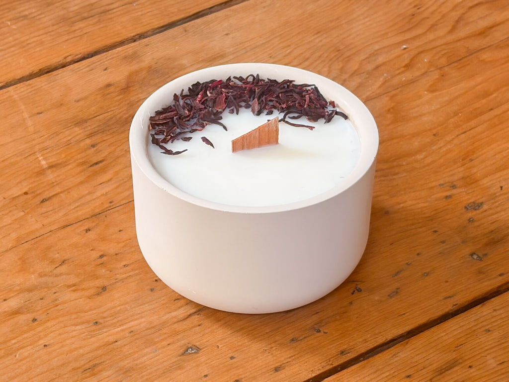 A white concrete candle holder filled with white wax, topped with dried dark red hibiscus flowers and a small wooden wick, set on a rustic wooden table.
