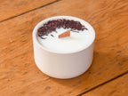 A white concrete candle holder filled with white wax, topped with dried dark red hibiscus flowers and a small wooden wick, set on a rustic wooden table.