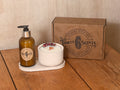 A Thirty 6 Scents luxury gift set on a wooden table, featuring an amber glass soap dispenser, a white jar candle with dried flowers on top, and a brown cardboard gift box.